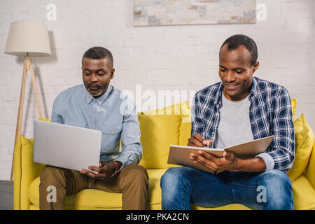 senior man using laptop and adult son writing in notebook at home Stock Photo