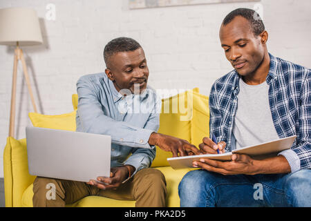 senior man using laptop and looking at adult son taking notes in notepad at home Stock Photo