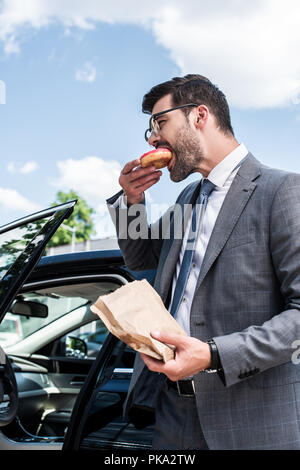 side view of businessman eating take away doughnut while standing at car on street Stock Photo