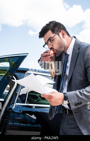 businessman eating doughnut and looking at documents while standing near car on street Stock Photo