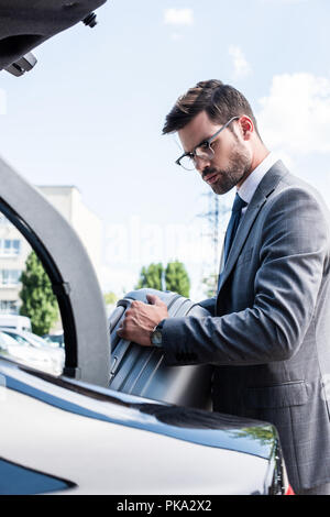 handsome businessman in eyeglasses putting wheeled bag in car trunk at street Stock Photo