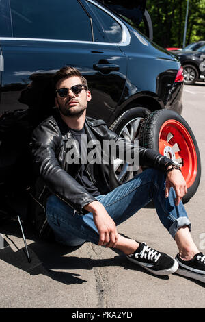 tired stylish man in sunglasses sitting near broken car at street Stock Photo