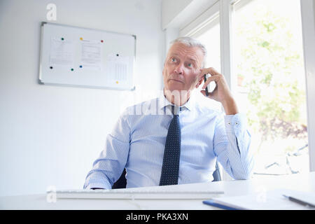 Senior businessman sitting at office desk and talking on his mobile phone. Elderly professional man wearing shirt and tie. Stock Photo