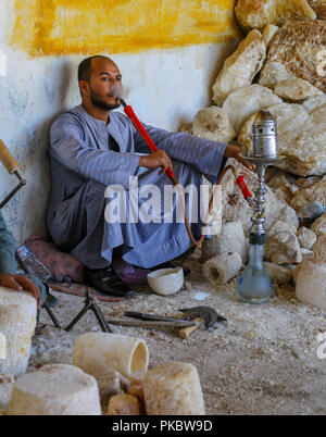 A worker at an Alabaster factory and shop having a break and smoking a Shisha pipe or Hooker pipe in Egypt, Africa Stock Photo