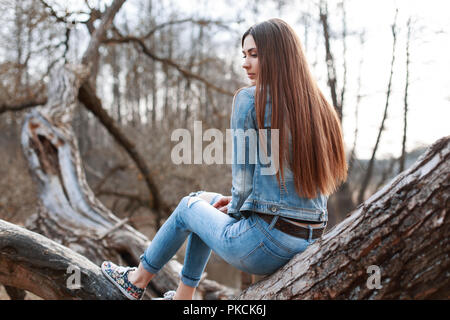 Young beautiful girl in denim jacket and jeans sits on a tree and dreams. Stock Photo