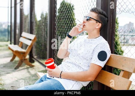 Young depressed man sitting on a bench and smokes. Guy holding a cup of coffee in his hand. Stock Photo
