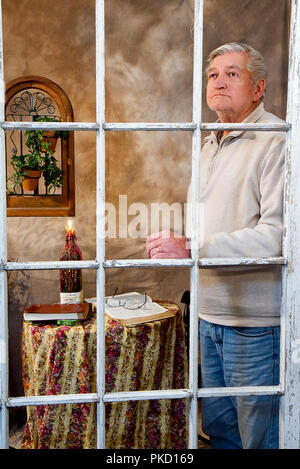 View through old worn window of a male senior citizen praying and/or looking out window with Bible and Candle on table in a retro designed room. Stock Photo