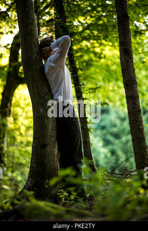 Young businessman in a suit standing leaning on a tree trunk under beautiful bright green tree crowns. Stock Photo