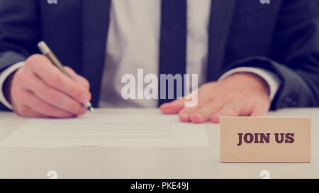 Join Us concept with a businessman sitting completing a job application or registration form with the words - Join Us on a wooden card in the foregrou Stock Photo