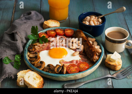 A traditional full English breakfast with fried egg, sausage, mushrooms, beans, bacon and tomatoes on a rustic wooden green table