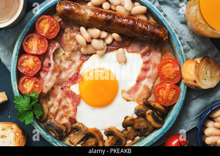 Traditional English breakfast close-up on a blue background, top view. Fried egg with sausage, mushrooms, beans, tomatoes and bacon.