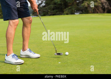 A mature male golfer's legs and footwear as he swings at the golf ball with a putter beside a marker on the putting green of a golf course