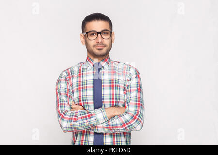 portrait of happy handsome bearded businessman in colorful checkered shirt, blue tie and black eyeglasses standing in crossed hands and smiling. indoo Stock Photo