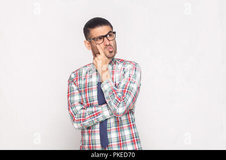 Portrait of confused handsome bearded businessman in colorful checkered shirt, blue tie and eyeglasses standing touching face and looking away. indoor Stock Photo