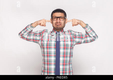 I don't want to hear. closed eyes unhappy bearded businessman in colorful checkered shirt, blue tie and eyeglasses standing with finger in ears. indoo Stock Photo