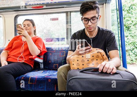 London England,UK,Piccadilly Circus Line,train inside interior man male woman female,sitting using smartphone earphones luggage riders passengers Stock Photo