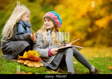 Cute little girls sketching outside on beautiful autumn day. Happy children playing in autumn park. Kids drawing with colourful pencils. Autumn activi Stock Photo