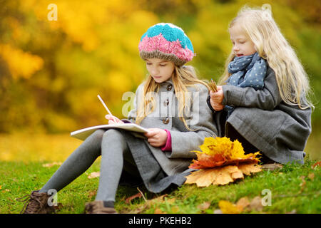 Cute little girls sketching outside on beautiful autumn day. Happy children playing in autumn park. Kids drawing with colourful pencils. Autumn activi Stock Photo