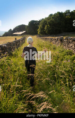 UK, Yorkshire, Wharfedale, Grassington, senior woman walking along footpath between drystone walls through traditional hay meadows Stock Photo