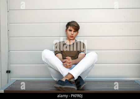 handsome young man in fashionable clothes is sitting on a bench near a metal wall Stock Photo