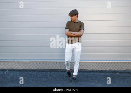 handsome model of a man in full growth in a fashion T-shirt with white pants and black shoes near the gray glossy wall Stock Photo
