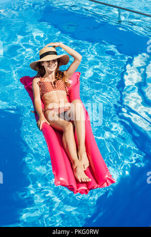 smiling young woman in straw hat and bikini  floating on inflatable mattress at swimming pool Stock Photo