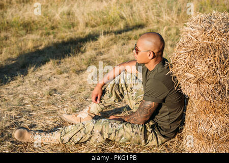 side view of young african american soldier in military uniform and sunglasses resting near hay on range Stock Photo