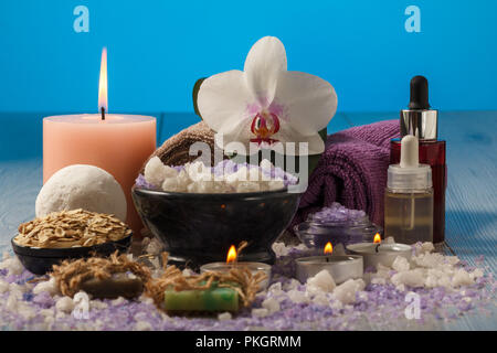 Spa composition with orchid flower, bowl with sea salt, bottles with aromatic oil, soap, scrub, candles and towels on wooden board and blue background Stock Photo