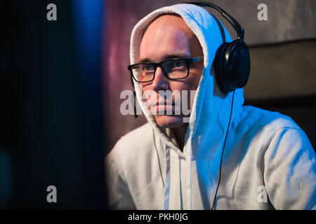 Portrait of concentrated young man in glasses and headset in dark room playing online computer game or competing in online esport tournament Stock Photo