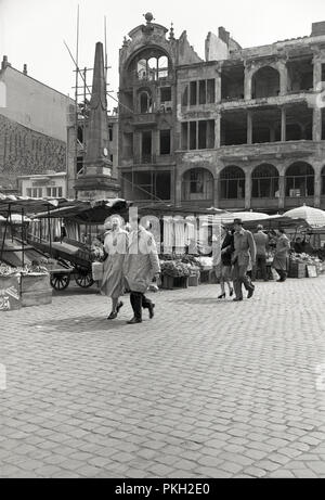 1950s, historical, people walking outside along a cobbled market square beside market stalls and traders, with destroyed buildings from WW2 seen in the background. Stock Photo