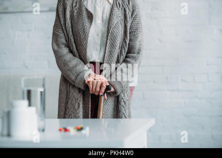 partial view of senior woman with walking stick standing in room with medicines on tabletop at home Stock Photo