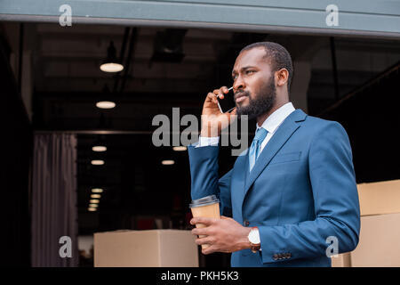 side view of african american businessman with coffee to go talking on smartphone