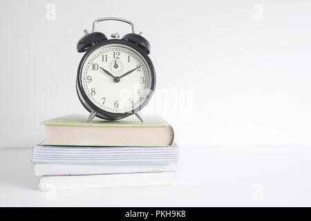 Alarm clock on a pile of books on a wooden shelf. White wall background and empty copy space for Editor's text. Stock Photo