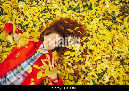 Autumn leaves falling on happy young woman in forest. Beautiful happy girl laying in yellow autumn leaves. Stock Photo