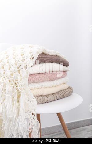 Stack of cozy knitted sweaters or blankets pastel trending colors on white modern chair against the wall Stock Photo