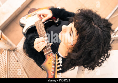 Top view portrait of a beautiful serious young woman in an elegant dress playing on an electric guitar while standing on a metal stairs. Stock Photo