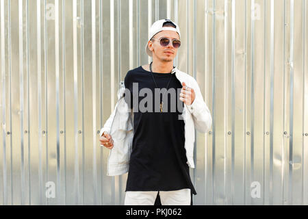 Stylish handsome man in a black T-shirt with a white jacket in fashion sunglasses near the metal wall Stock Photo