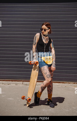 stylish tattooed woman in sunglasses listening music with earphones and standing near skateboard Stock Photo