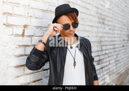Fashionable young handsome man wearing sunglasses, a black hat and stylish fashion jacket near vintage white brick wall Stock Photo