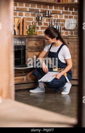 serious young repairwoman with clipboard sitting near oven and looking at it Stock Photo