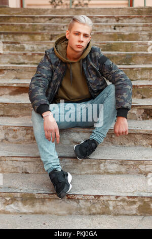 Stylish young man in a military jacket with a hoodie and jeans posing on the steps Stock Photo