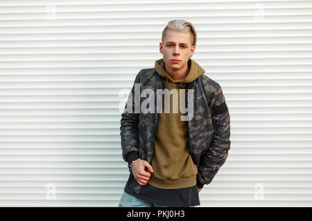 Handsome young man in stylish military jacket with a hoodie near white metal walls Stock Photo