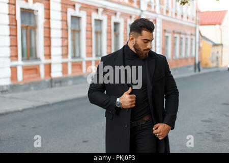 handsome stylish strong man in a black trendy coat walks on the street Stock Photo