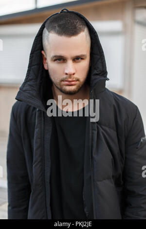 Stylish beautiful young man with a hood in a black winter jacket on the street Stock Photo