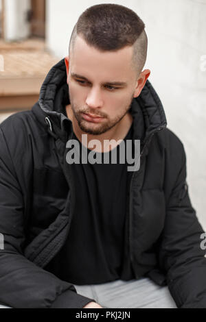 Portrait of a young man with a short hair in a warm winter jacket sits on the stairs Stock Photo