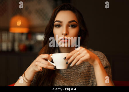 Young beautiful woman in a knitted sweater drinks delicious coffee in a restaurant Stock Photo