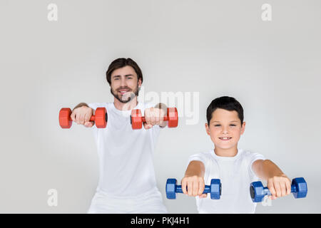 portrait of smiling father and son in white shirts exercising with dumbbells on grey background Stock Photo