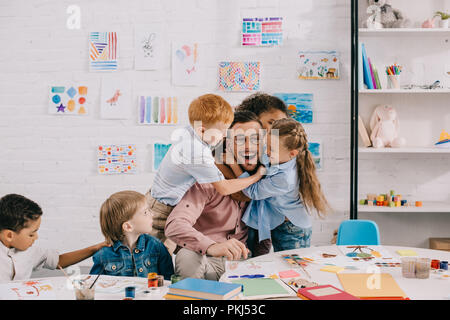 interracial kids hugging happy teacher at table in classroom Stock Photo