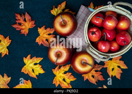 Apples in a straw basket over a black background of maple leaves scattered in the scene. Concept for fall and autumn Stock Photo
