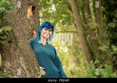 Portraits of a blue haired woman, in a blue jumper, leaning against a tree trunk Stock Photo